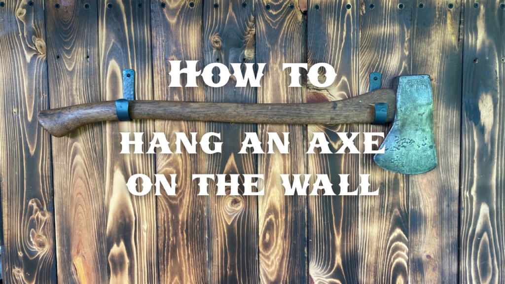 A photo of a Plumb brand axe mounted on a charred pine backdrop using two blacksmith made wall hooks with a text overlay that says "How to Hang an Axe on the wall"